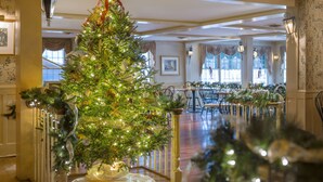 Un amplio vestíbulo de hotel con un gran árbol de Navidad decorado con luces y adornos, un comedor con mesas dispuestas para una comida, y elegantes decoraciones en las paredes.