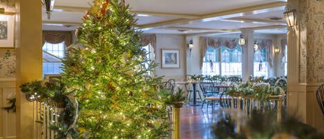 A grand hotel lobby with a large Christmas tree adorned with lights and ornaments, a dining area with tables set for a meal, and elegant wall decorations.