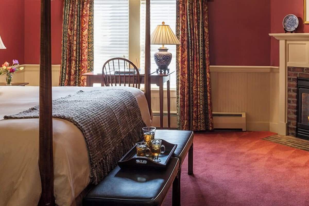 A hotel room with a four-poster bed, a fireplace, a dining table with a tray of drinks, and a window with curtains.
