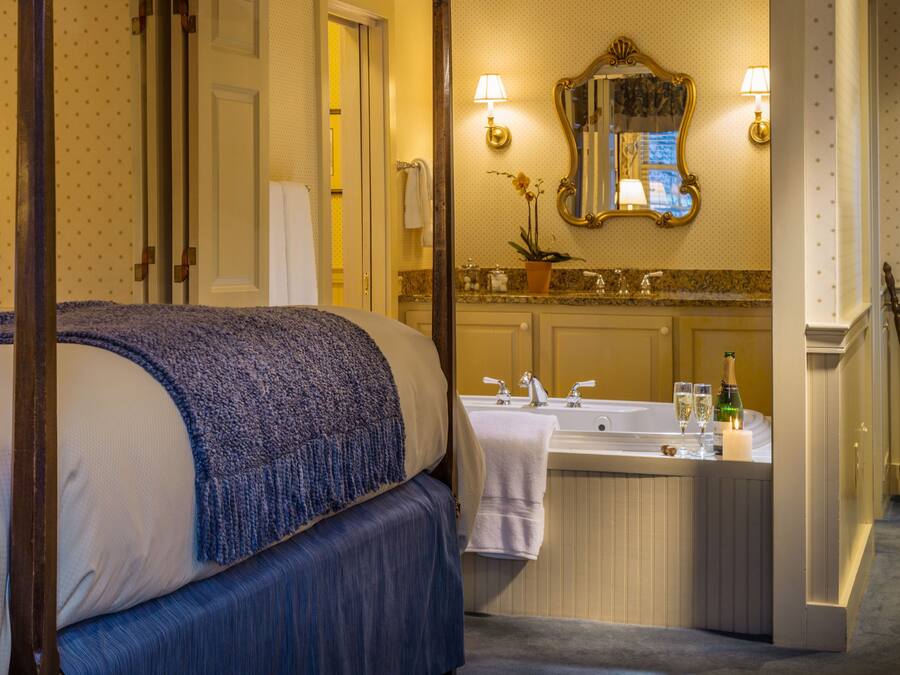 Quarto de hotel com uma cama grande, banheiro com pia e espelho, e uma porta que dá para outro cômodo.