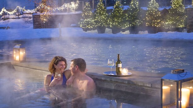 Zona de piscina al aire libre climatizada con una pareja disfrutando del calor, rodeada de nieve y luces navideñas.