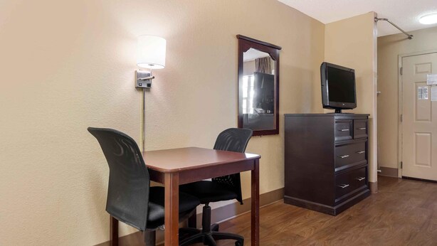 A hotel room with a wooden desk, two black office chairs, a dark dresser, a flat-screen TV, and a wall-mounted mirror.