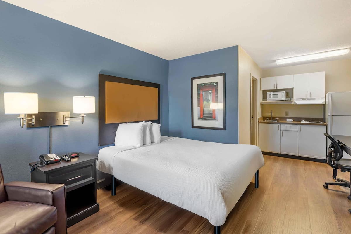 A hotel room with a bed, a desk with a lamp, a chair, a kitchenette, and a framed picture on the wall.