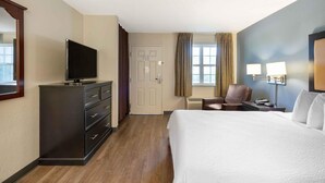 A hotel room with a bed, a TV on a dresser, a desk with a chair, and a window with curtains.