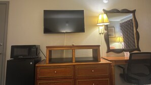 A hotel room with a flat-screen TV mounted on the wall, a wooden dresser with drawers, a microwave on top of a refrigerator, a desk with a chair, and a lamp. - Days Inn by Wyndham Jacksonville (Jacksonville)