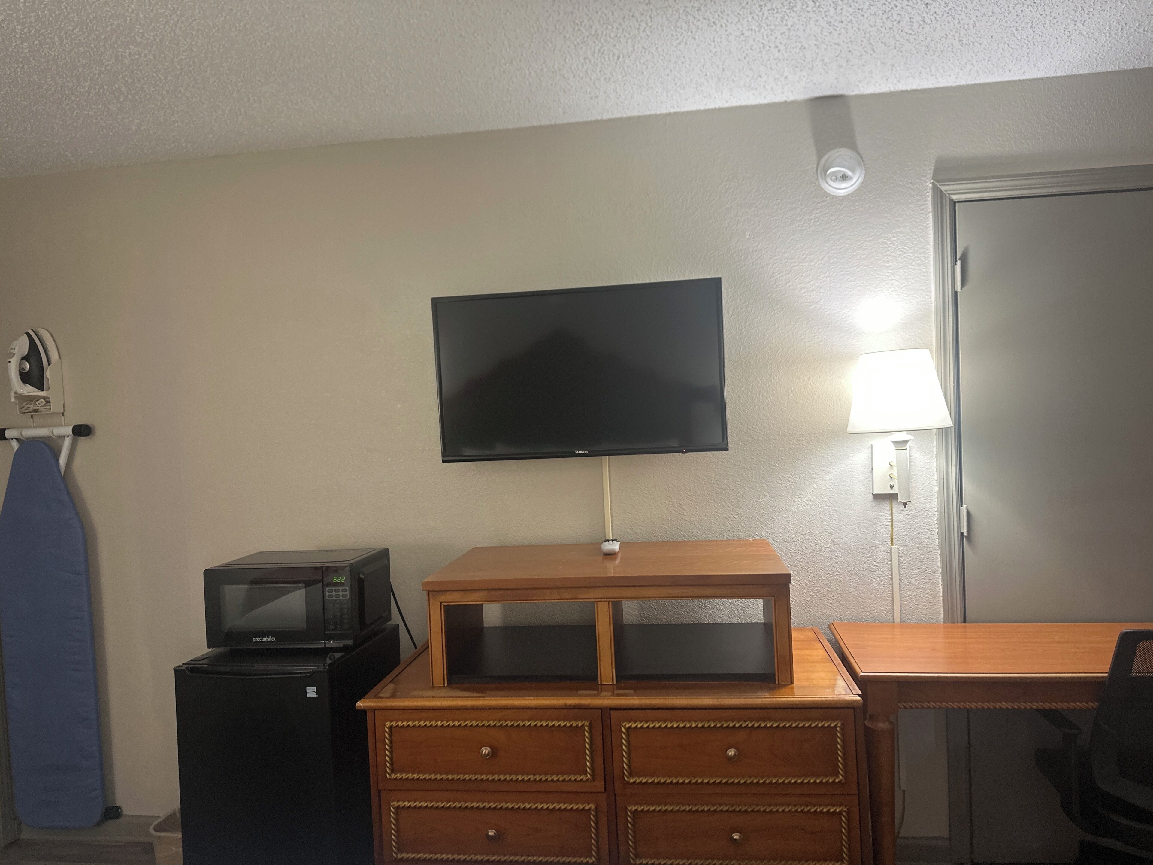A room with a television mounted on the wall, a wooden dresser, a microwave, and a lamp.