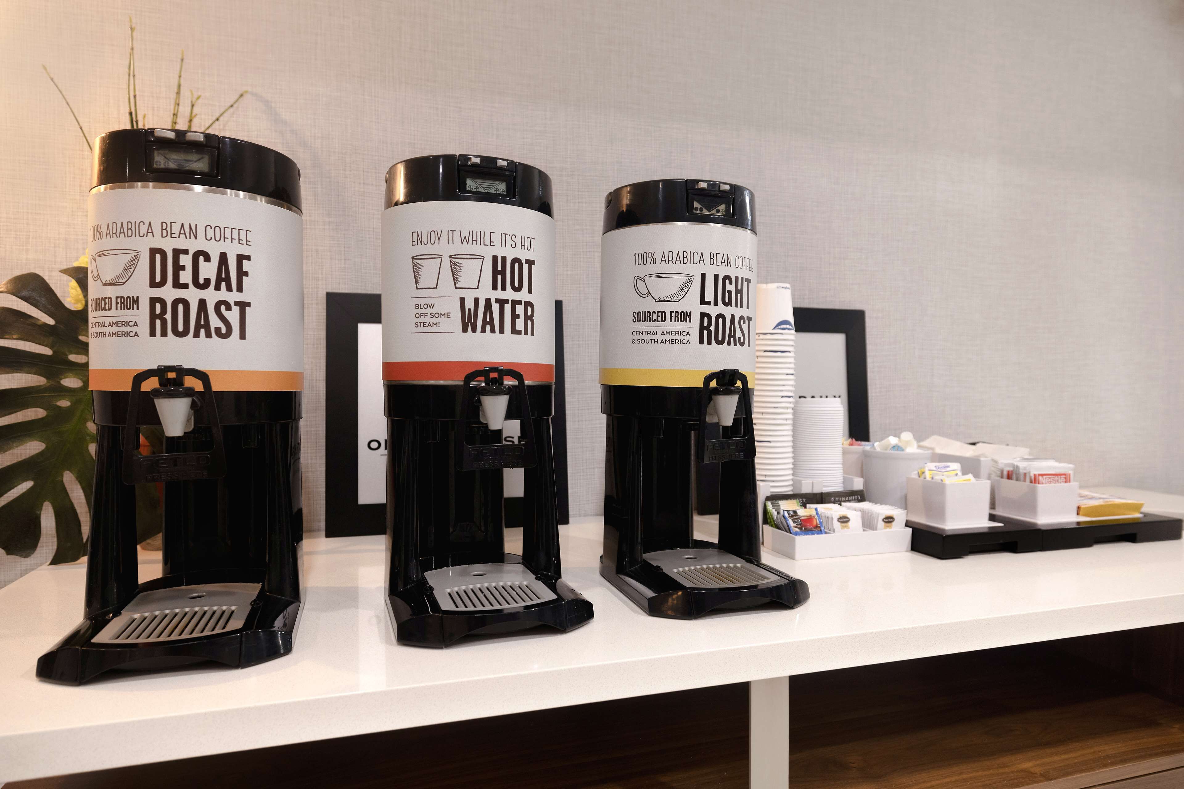 Tres dispensadores de café, etiquetados como 'Decaf Roast', 'Agua Caliente' y 'Light Roast', con cápsulas de café y tazas sobre una encimera.