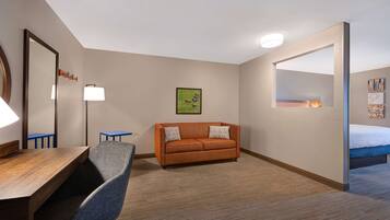 Habitación de hotel con un escritorio de madera, una silla azul, un sofá naranja, una cama, un espejo grande y un cuadro en la pared.