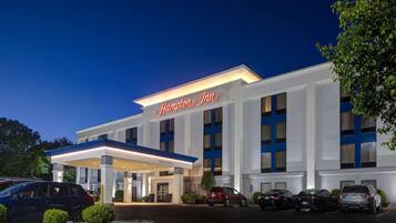 Un hotel Hampton Inn con una fachada moderna, una entrada techada y un estacionamiento con varios coches.