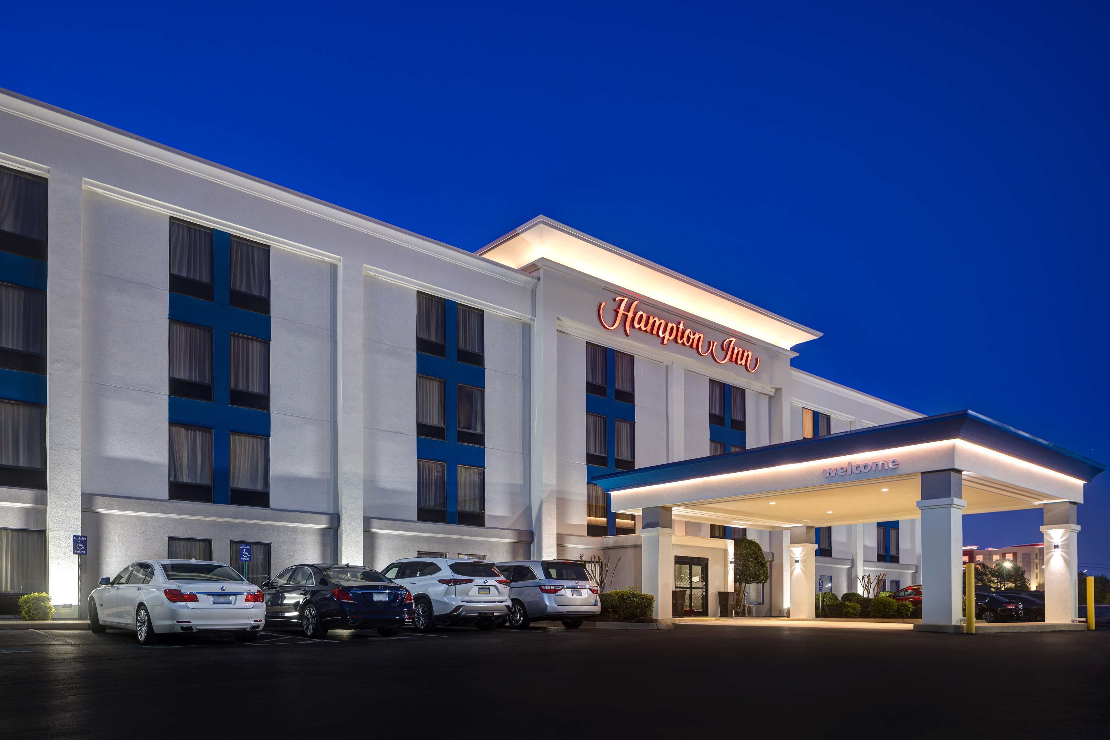 Un hotel Hampton Inn con una fachada moderna, un acceso cubierto y varios coches estacionados delante.