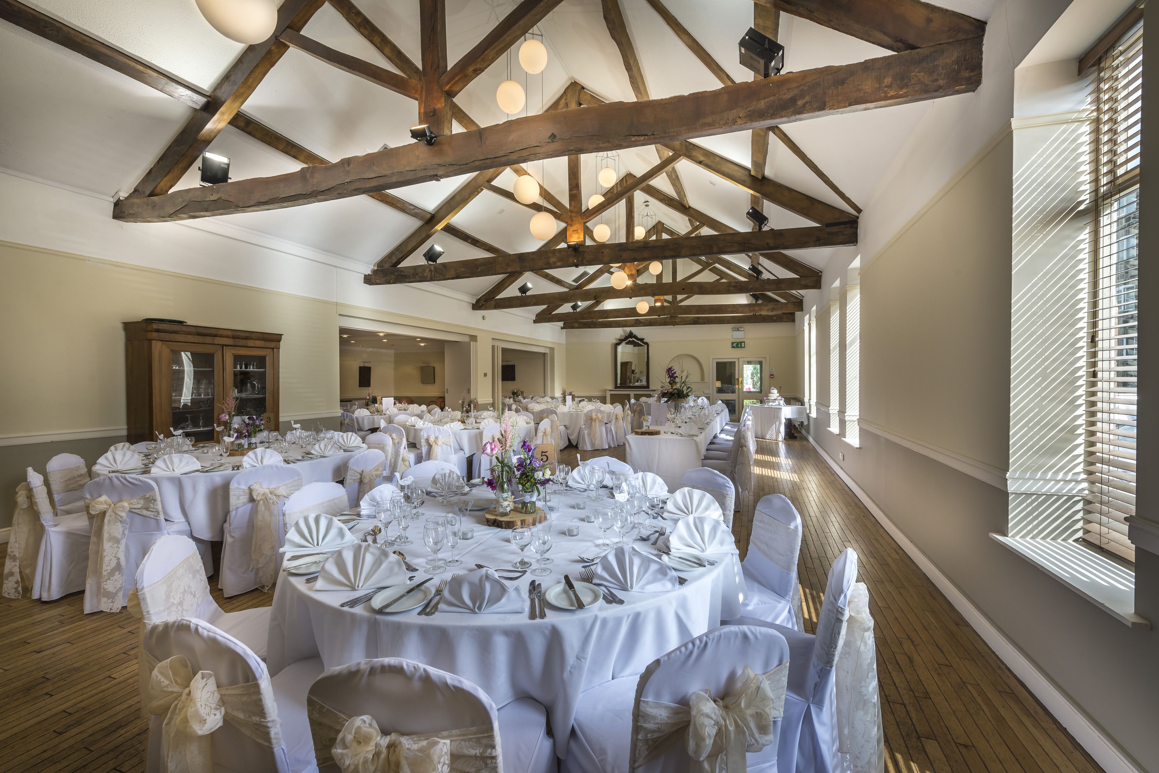 A banquet hall with round tables set for a formal event, wooden beams on the ceiling, and large windows on one side.