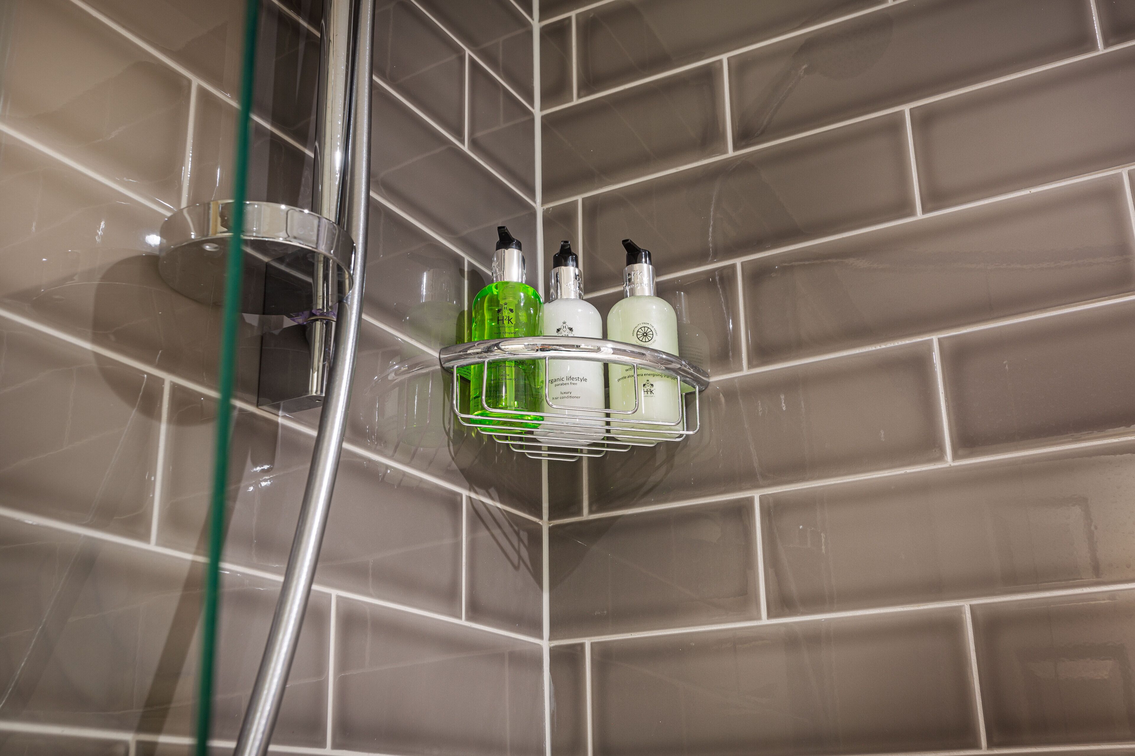 A bathroom corner with a shower, a glass shelf holding toiletries, and a tiled wall.
