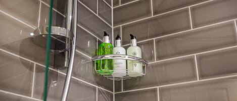 A bathroom corner with a shower, a glass shelf holding toiletries, and a tiled wall.