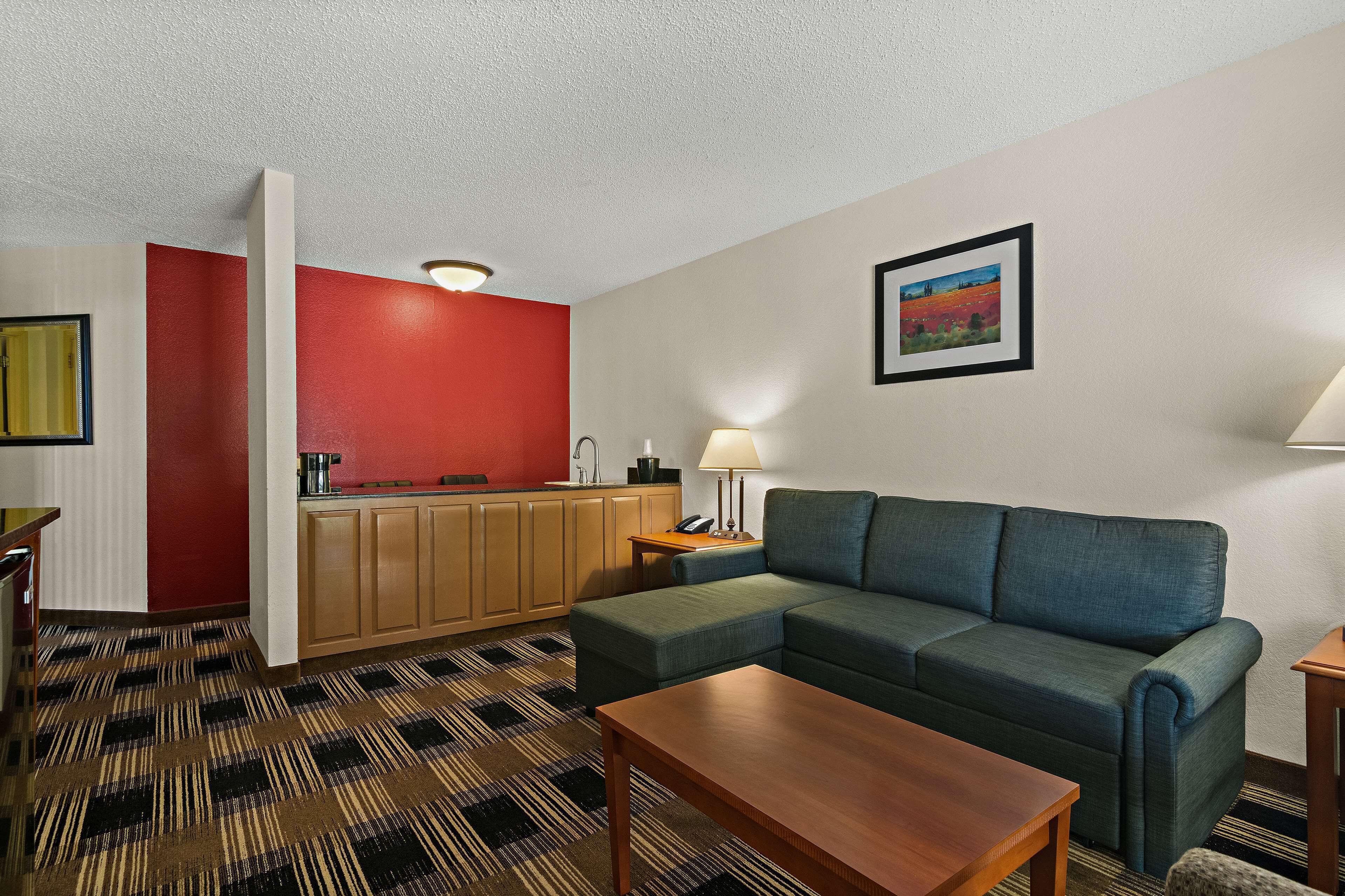 Hotelkamer met een groene bank, een houten salontafel, een keukenhoek met rode accentmuur, en een schilderij aan de wand.