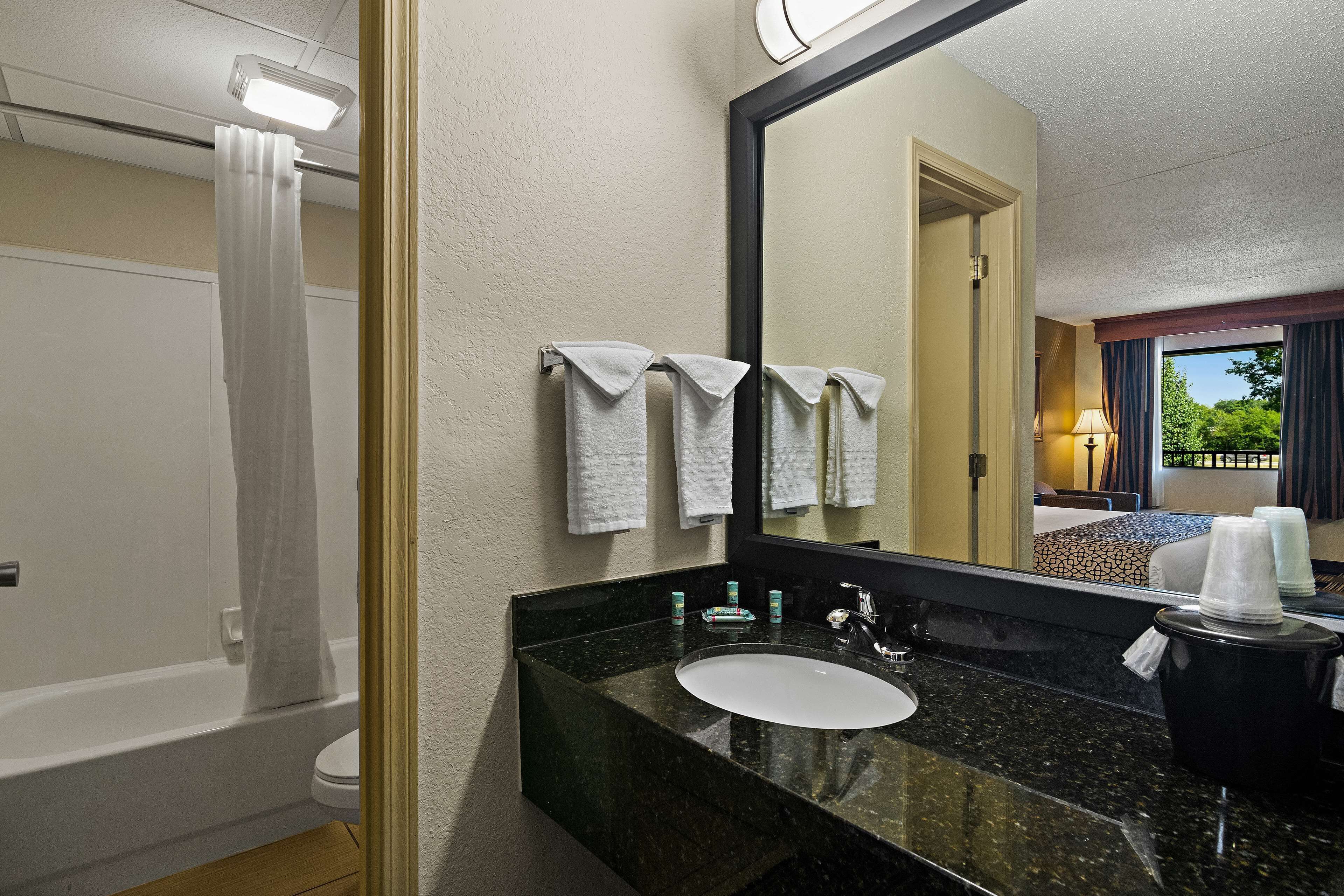 A hotel bathroom with a bathtub, a sink, and a large mirror. There are towels hanging on the wall and a window with curtains.