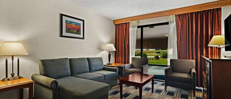 Ein Hotelzimmer mit einer Couch, einem Sessel und einem kleinen Tisch. Ein Fenster mit Vorhängen bietet Blick auf eine Landschaft.
