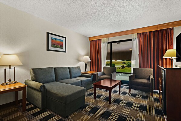 A hotel room with a sofa, armchair, and a small table. There is a window with curtains and a view of a landscape.