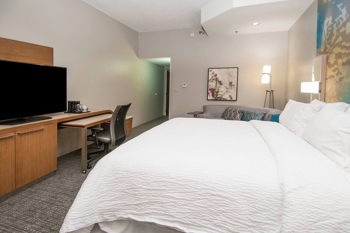 Ein Hotelzimmer mit einem Bett, einem Schreibtisch mit Flachbildfernseher, einem Sessel, einer Lampe und einem Bild an der Wand.