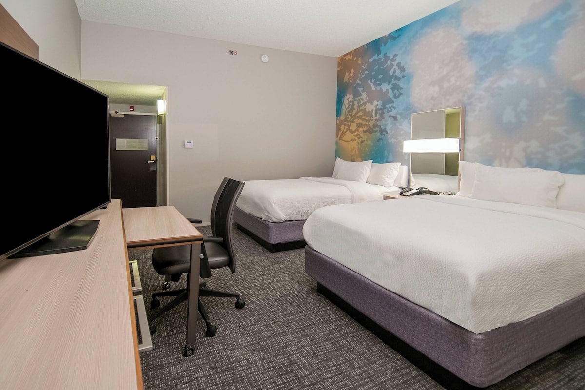 Ein Hotelzimmer mit zwei Betten, einem Schreibtisch mit Computer, einem Fernseher und einem großen Fenster mit Blick auf Bäume.