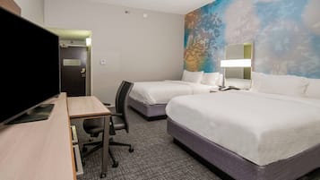 Une chambre d’hôtel avec deux lits, un bureau avec un ordinateur, une télévision et une grande fenêtre donnant sur des arbres.
