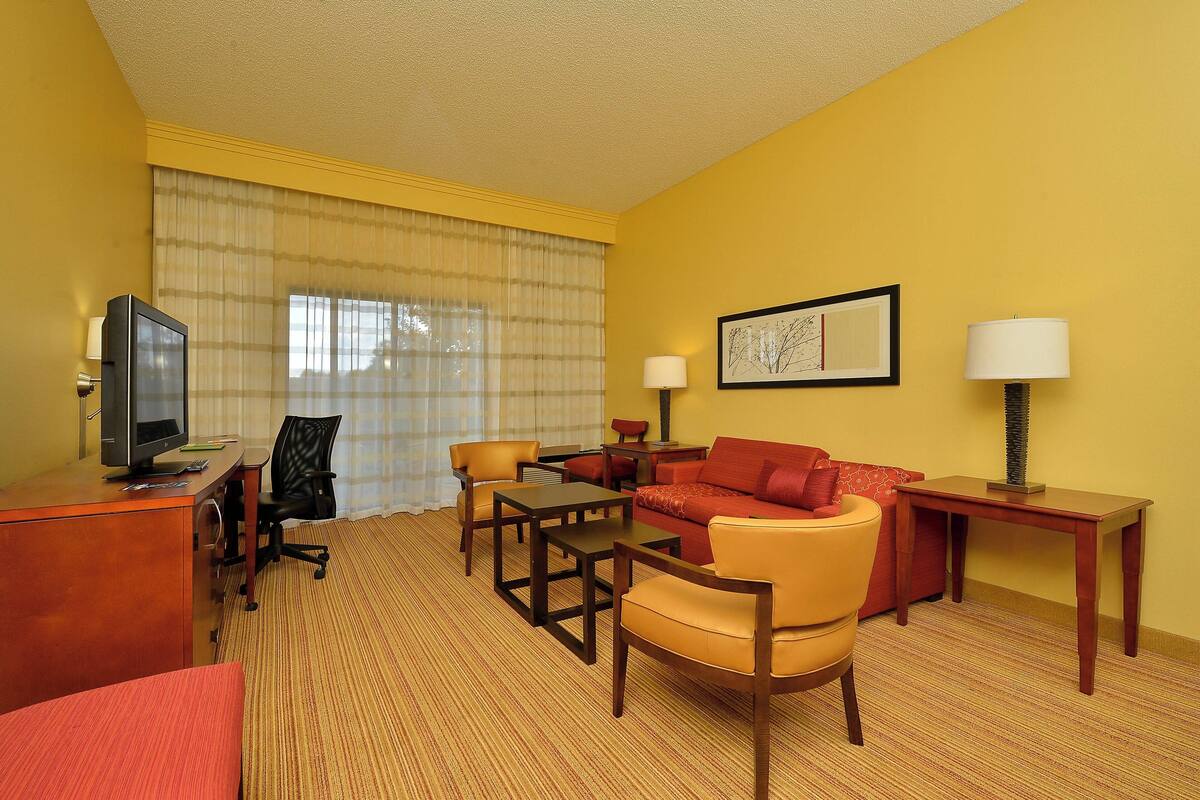Ein Hotelzimmer mit gelber Wand, einem großen Fenster mit transparenten Vorhängen, einer roten Couch, einem Holztisch mit Fernseher, einer Lampe und einem gerahmten Bild.