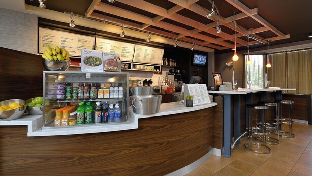 A bar area with a wooden counter, a refrigerated display with beverages, and a menu board. - Courtyard By Marriott Bentonville (Bentonville)