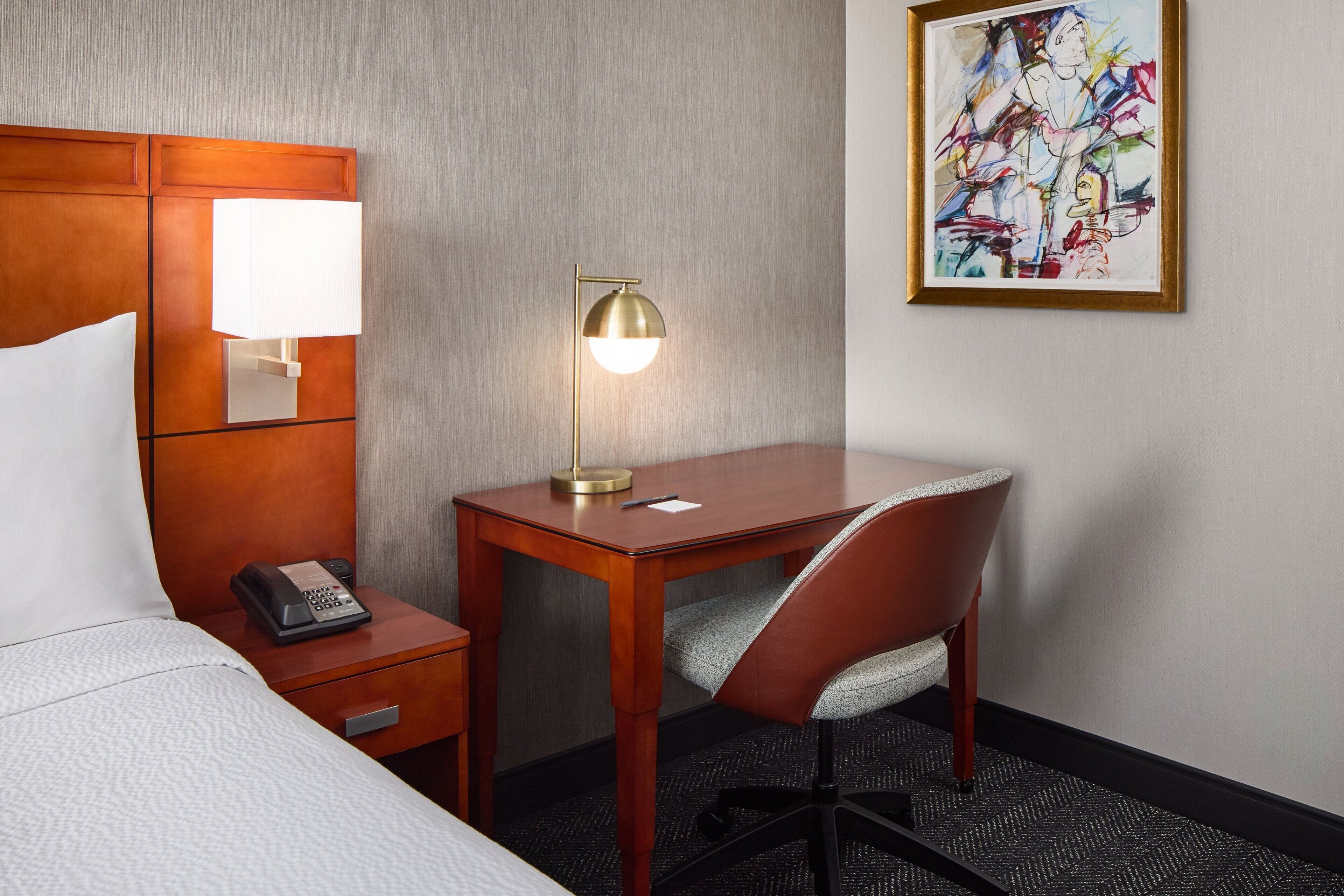 A hotel room with a bed, a desk with a lamp, a chair, a telephone, and a painting on the wall.