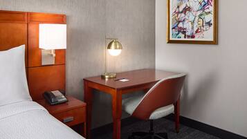 A hotel room with a bed, a desk with a lamp, a chair, a telephone, and a painting on the wall.
