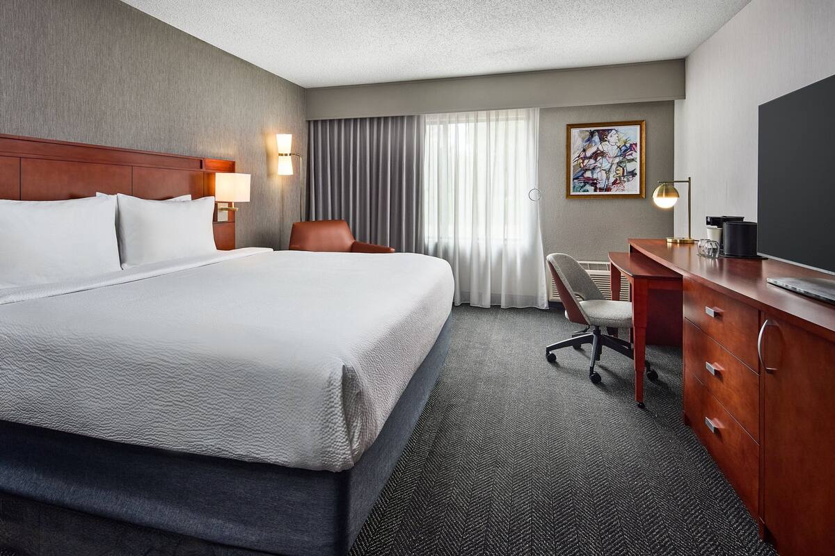 A hotel room with a large bed, a desk with drawers, a chair, a lamp, and a painting on the wall.