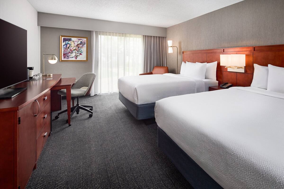 A hotel room with a large bed, a smaller bed, a desk with a chair, a TV, and a painting on the wall.