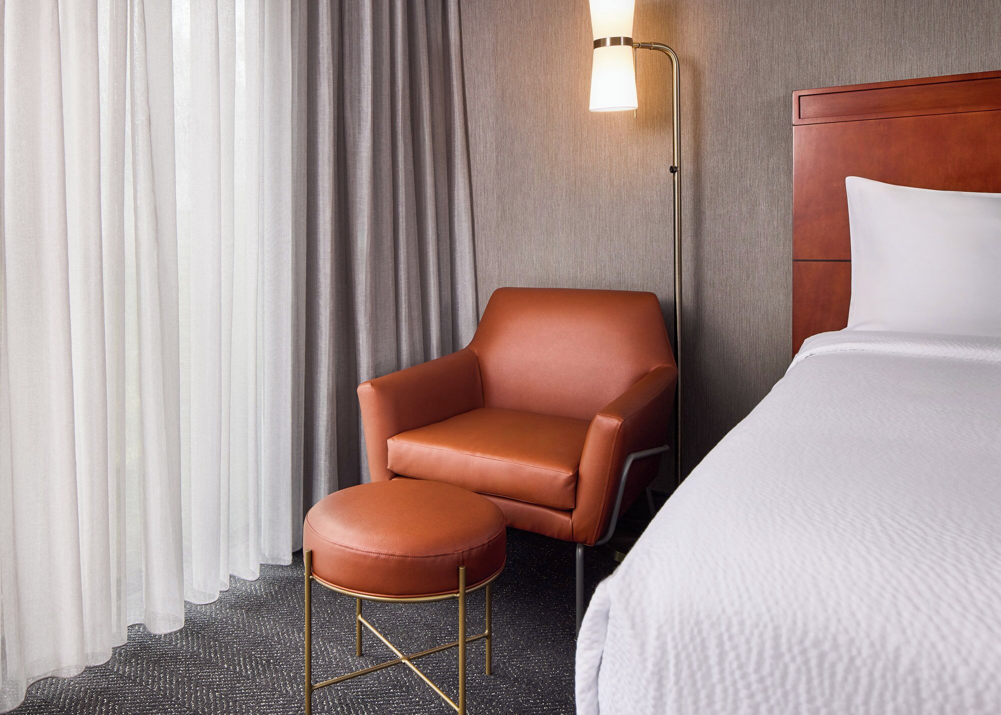 Ein Hotelzimmer mit einem Bett, einem braunen Ledersessel, einem kleinen Hocker, einer Stehlampe und einem Fenster mit Vorhängen.