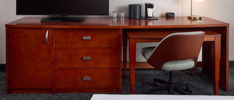 A hotel room with a wooden desk, a flat-screen TV, a coffee maker, a lamp, and a chair.
