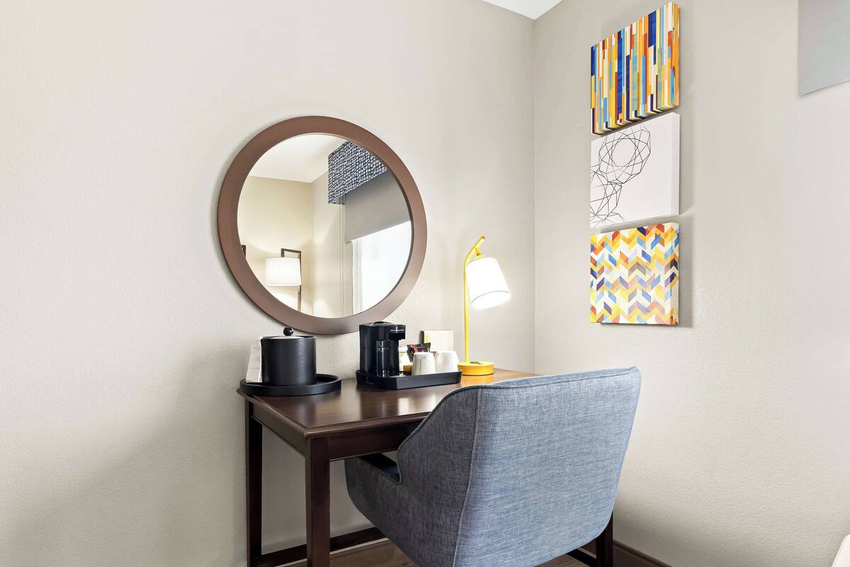 A hotel room with a desk, a round mirror, a coffee maker, a lamp, and a chair.