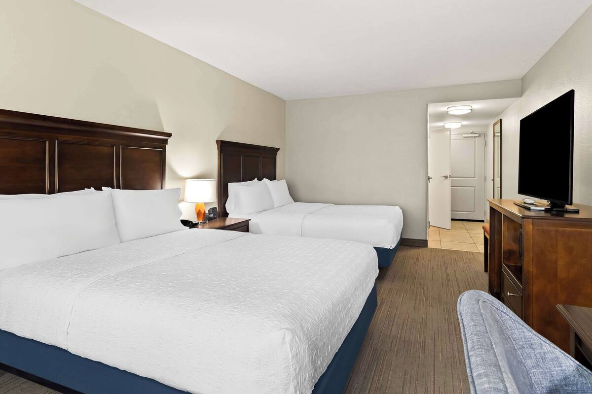 A hotel room with two beds, a desk, a television, and a door leading to another room.