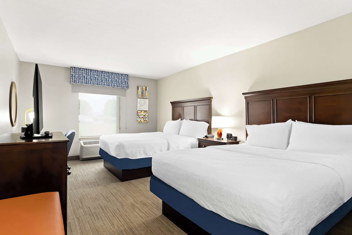 A hotel room with two beds, a desk, a chair, a TV, and artwork on the wall.