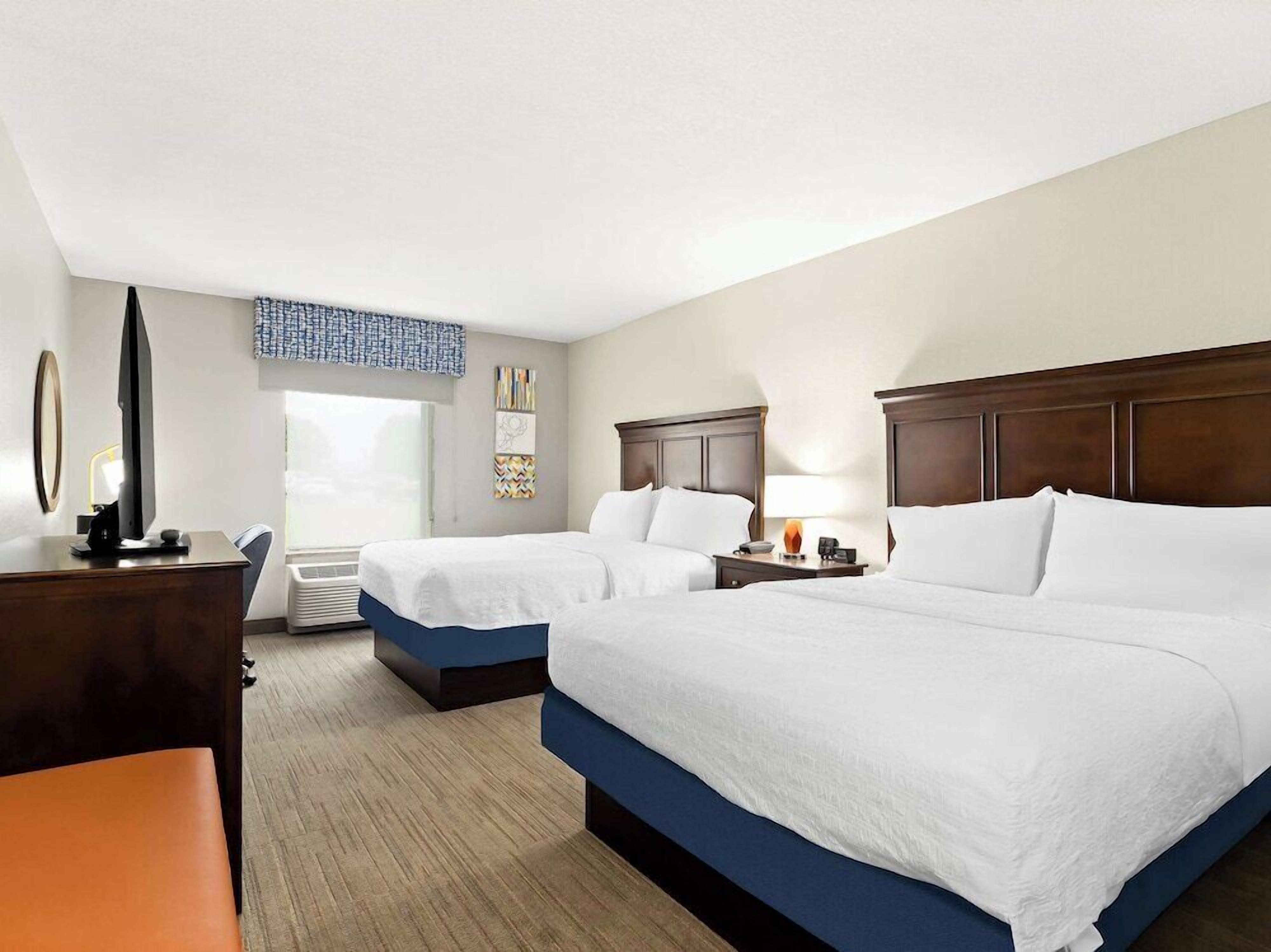 A hotel room with two beds, a desk, a chair, a TV, and a window.