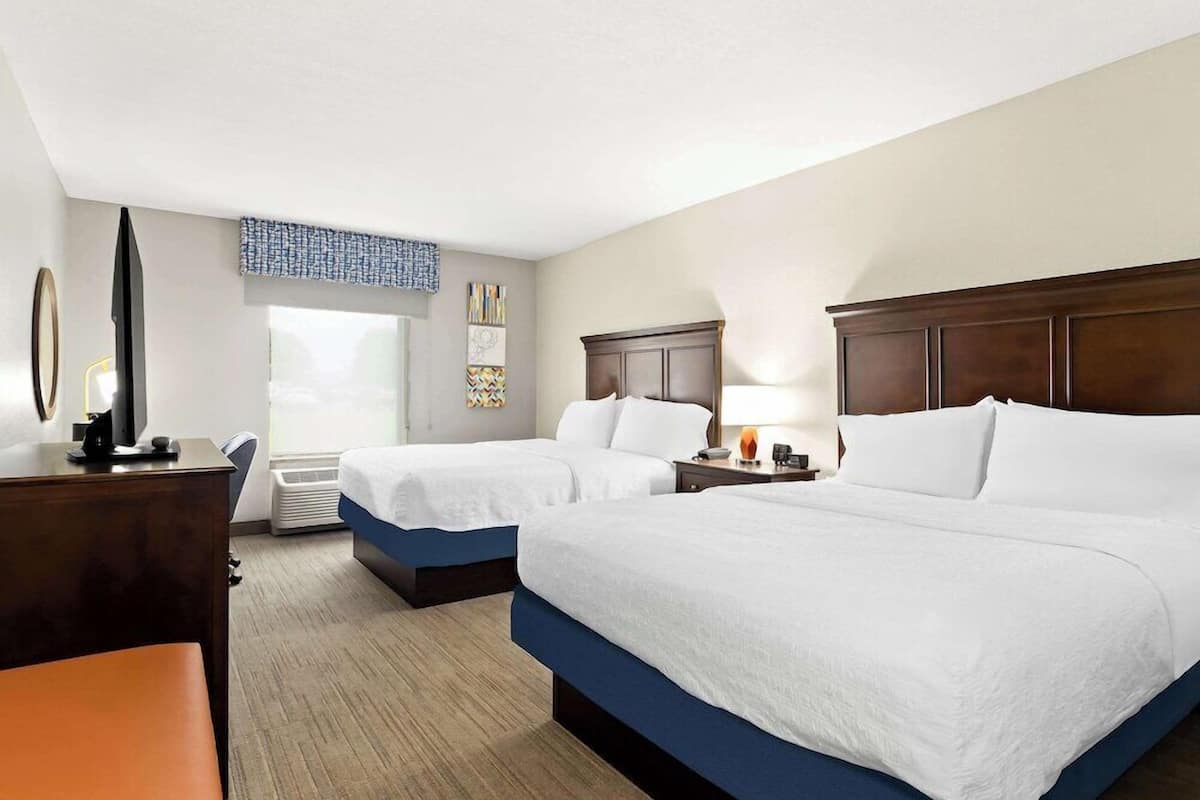 A hotel room with two beds, a desk, a chair, a TV, and a window.