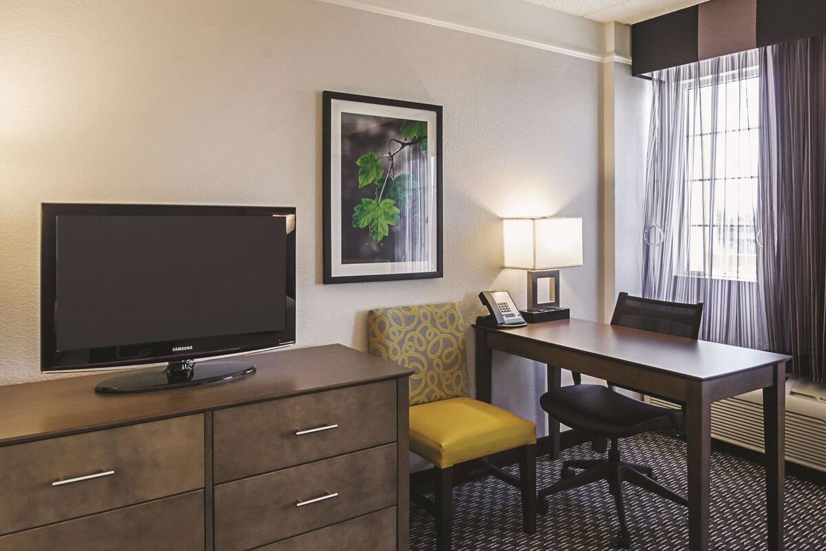 A hotel room with a flat-screen TV, a desk with a lamp, a chair, a sofa, and a framed picture on the wall.