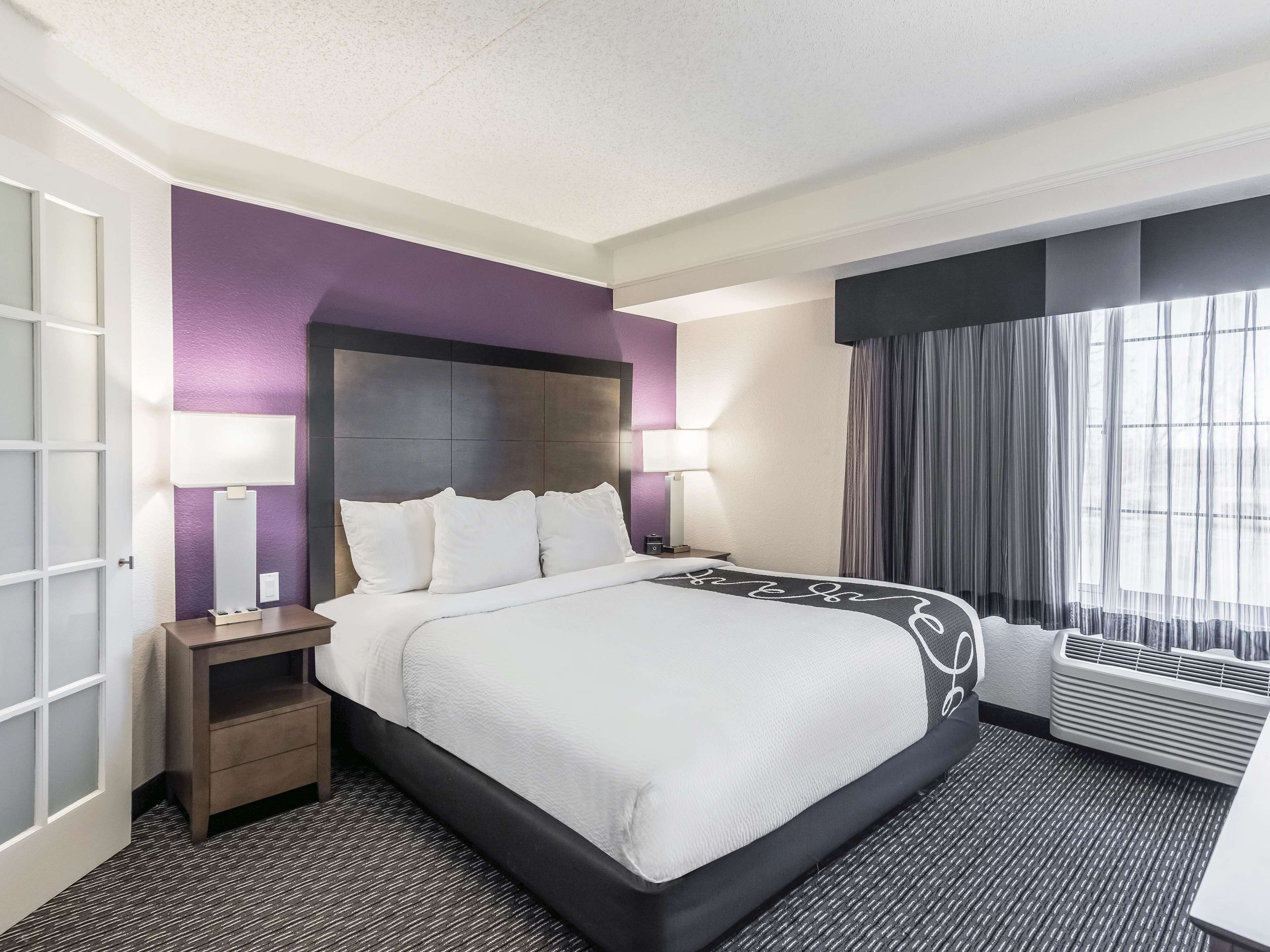 A hotel room with a large bed, two bedside tables, a window with curtains, and a wall-mounted air conditioning unit.