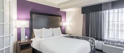 A hotel room with a large bed, two bedside tables, a window with curtains, and a wall-mounted air conditioning unit.