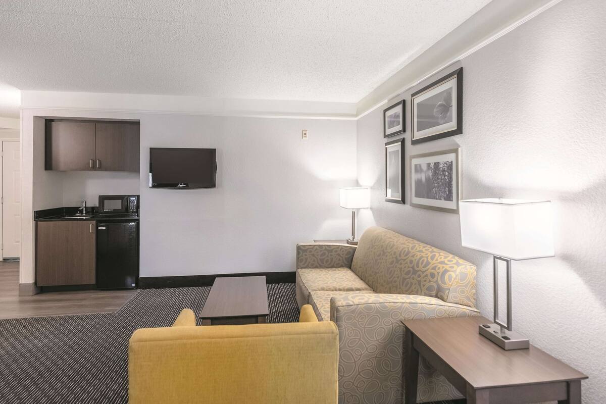 A modern hotel room with a kitchenette, a sofa, a small table, and a TV mounted on the wall.