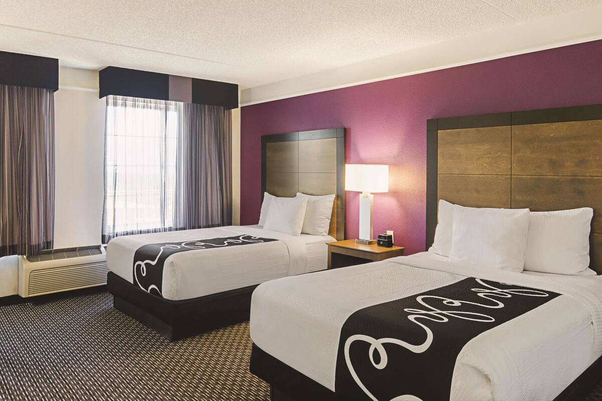 A hotel room with two beds, a nightstand with a lamp, a window with curtains, and a wall heater.