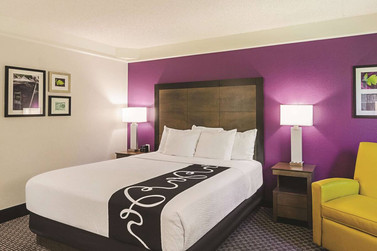 A hotel room with a bed, two bedside lamps, a nightstand, a chair, and framed pictures on the wall.