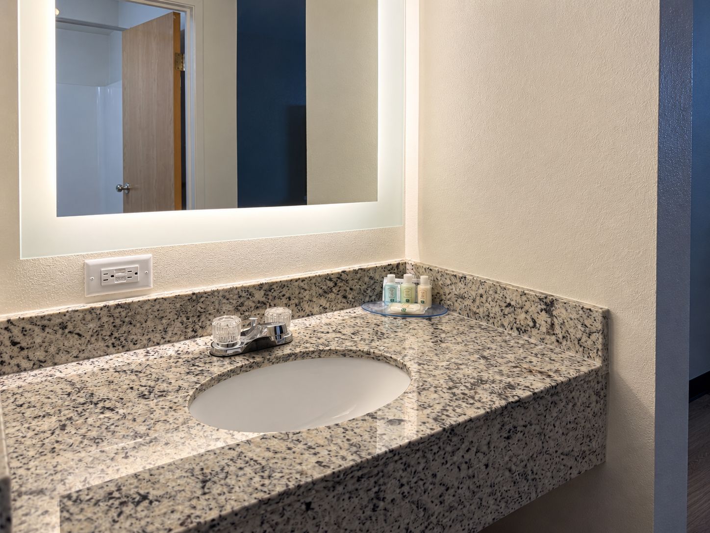 A bathroom with a granite countertop, a sink, and a mirror. There are toiletries on the countertop.