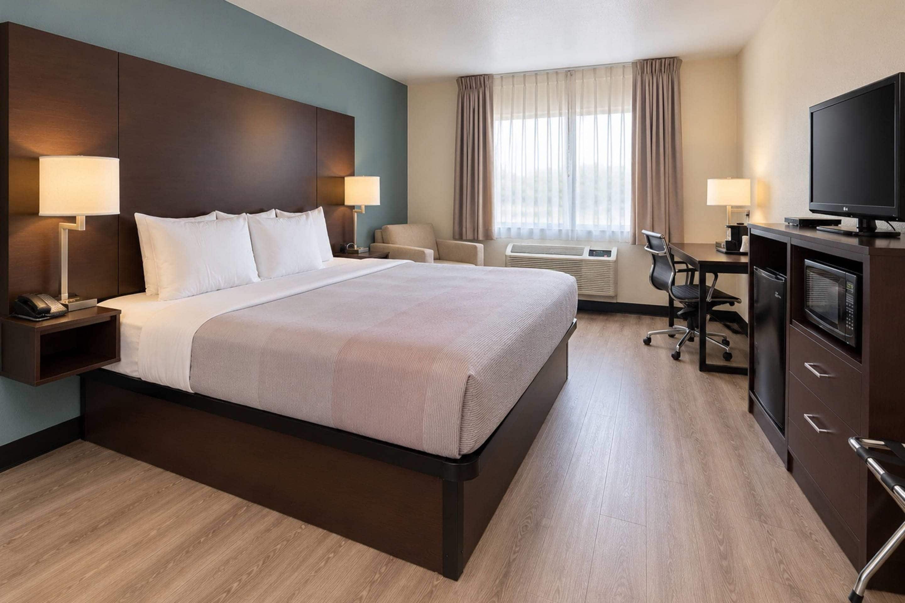 A hotel room with a large bed, bedside lamps, a desk with a chair, a television, and an air conditioning unit.