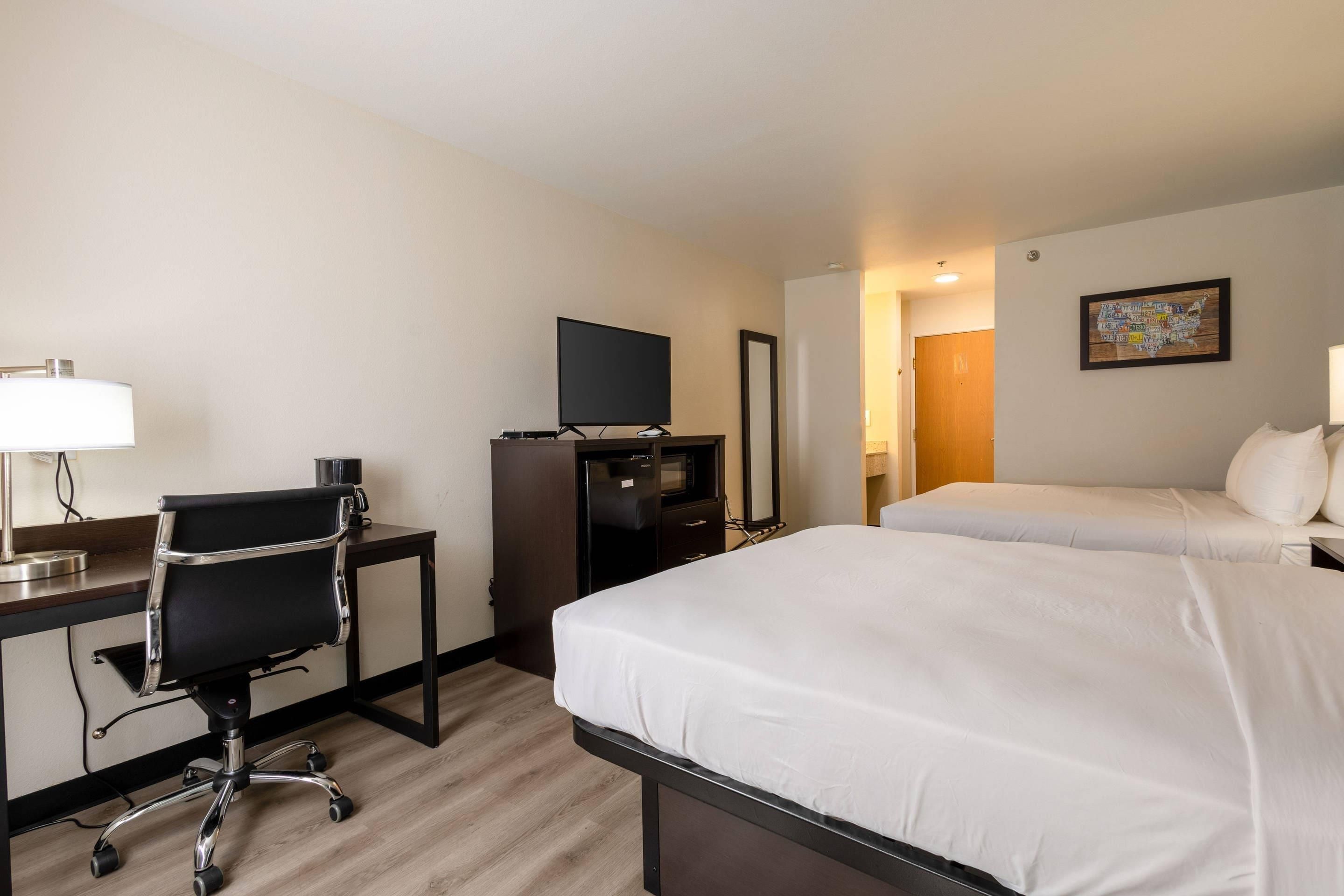 Habitación de hotel con escritorio, silla, dos camas, refrigerador, televisor y espejo montado en la pared.