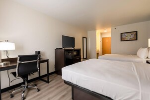 Habitación de hotel con escritorio, silla, dos camas, refrigerador, televisor y espejo montado en la pared.