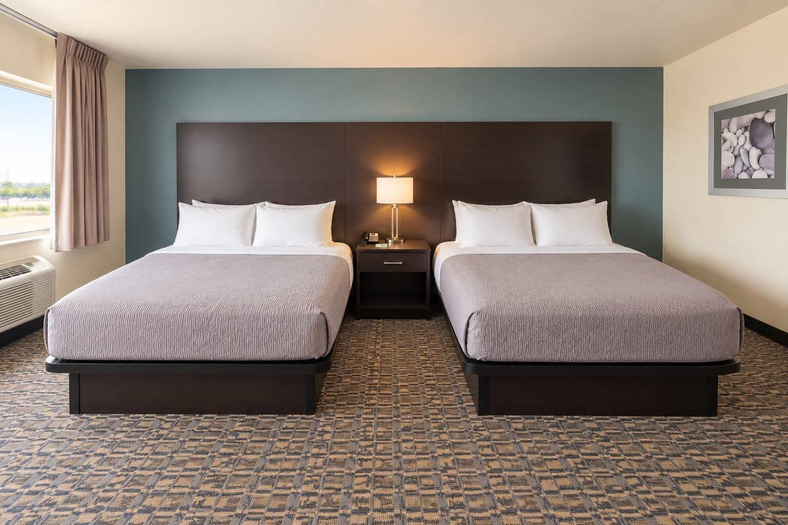 Habitación de hotel con dos camas, una mesita de noche con lámpara, una ventana con cortinas y una alfombra estampada.