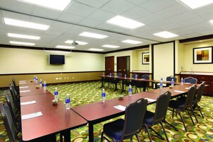 Un salón de conferencias con mesas largas, sillas, botellas de agua, un proyector y una televisión en la pared.