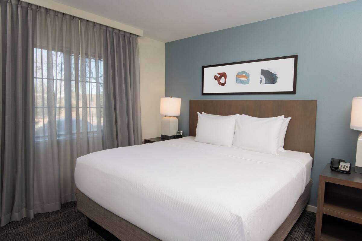 A hotel room with a large bed, two bedside tables with lamps, a framed artwork on the wall, and a window with curtains.